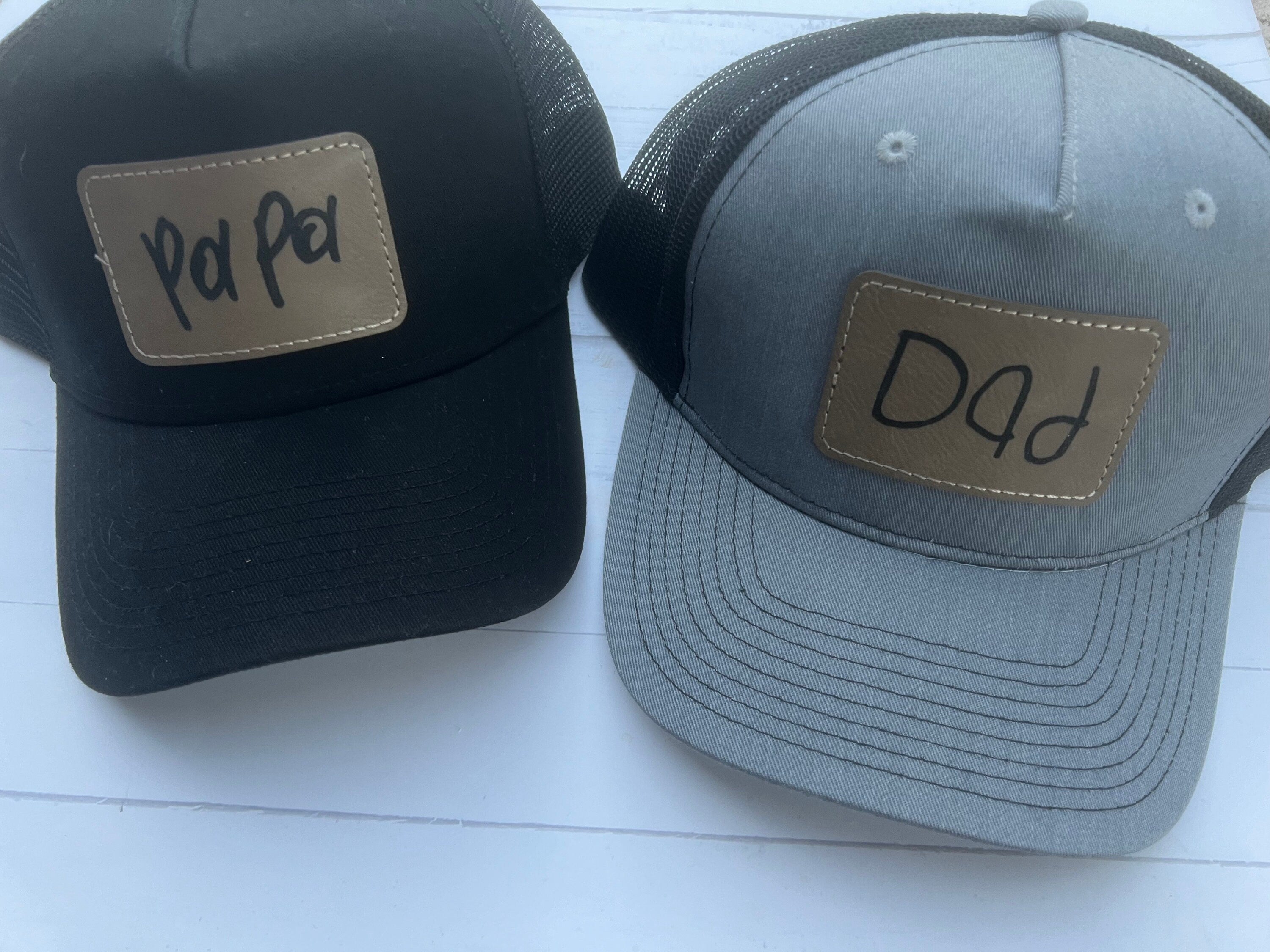 May include: Two black and gray trucker hats with gold patches. The left hat has a gold patch with the word 'Pa Pa' written in black. The right hat has a gold patch with the word 'Dad' written in black.