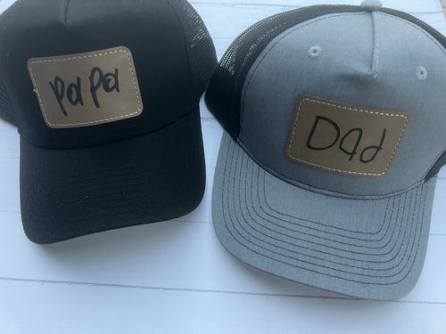 May include: Two black and gray trucker hats with gold patches. The left hat has a gold patch with the word 'Pa Pa' written in black. The right hat has a gold patch with the word 'Dad' written in black.